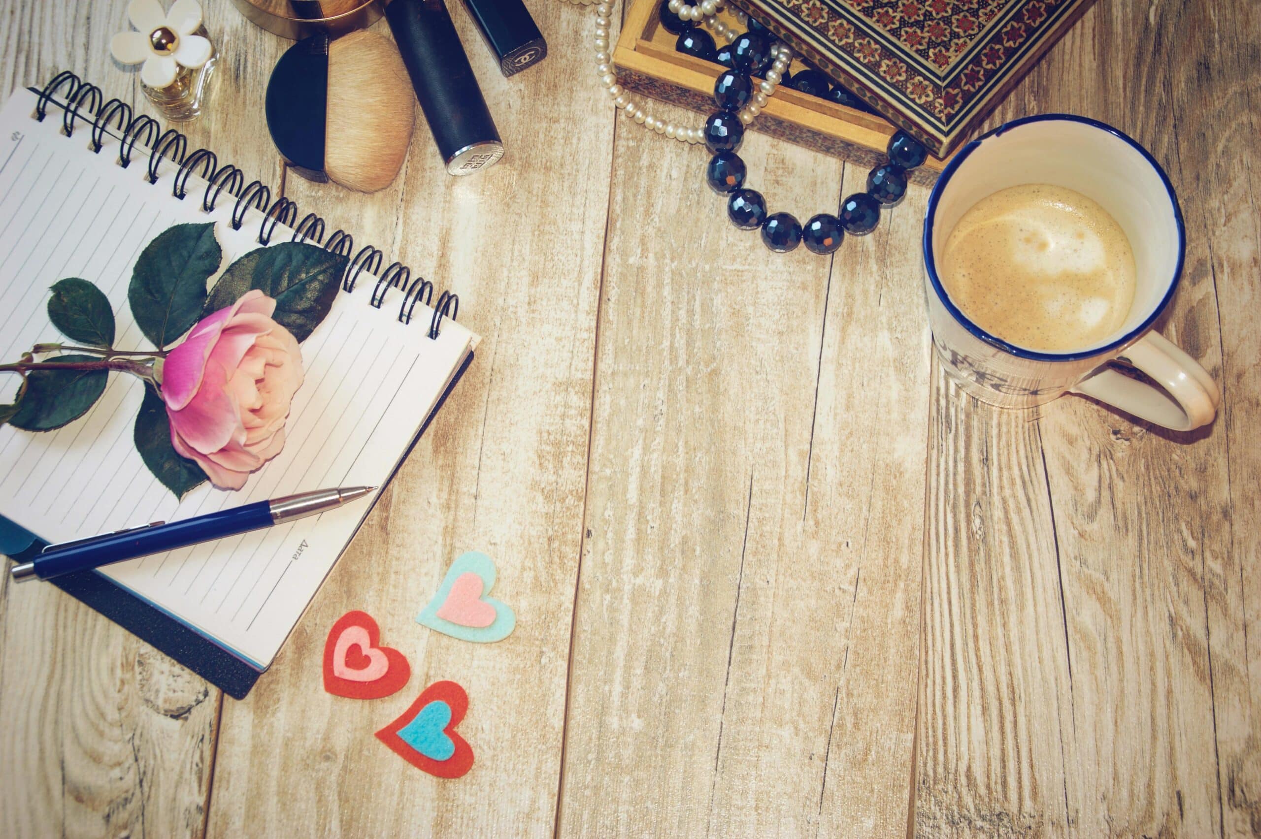 Flat lay of coffee, notebook, rose, and accessories on wooden table.